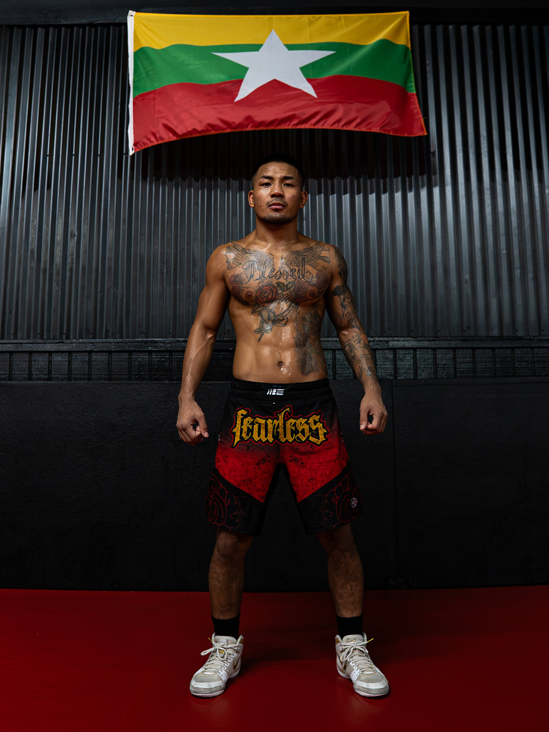 A shirtless tattooed man stands in a gym wearing red and black fight shorts with 'Fearless' printed in yellow across the front, black socks, and white sneakers. A Myanmar flag hangs on the dark corrugated wall behind him, and a red mat covers the floor.